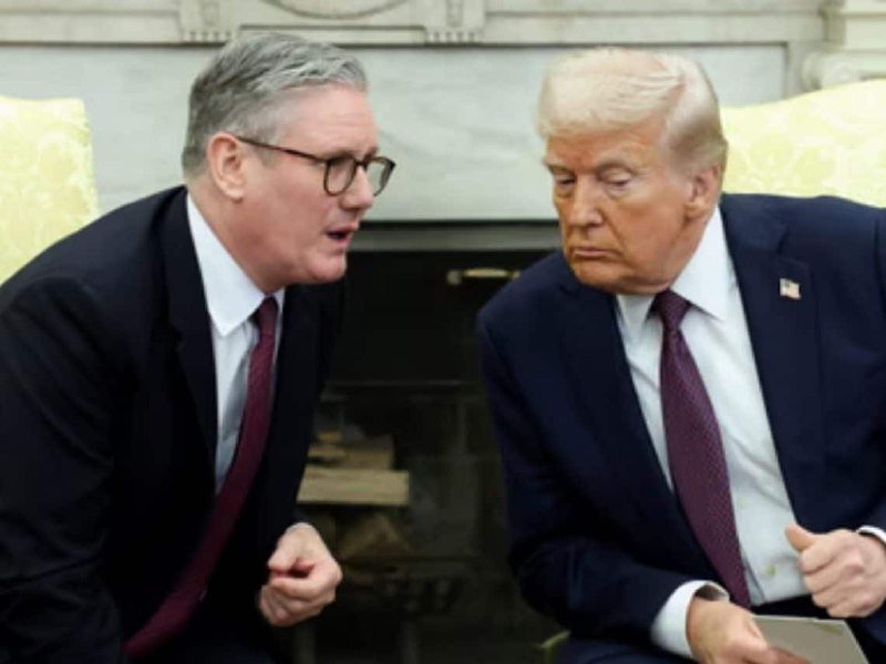 Two men in dark suits sit side by side in front of a fireplace, one wearing glasses and speaking while the other listens with a serious expression.