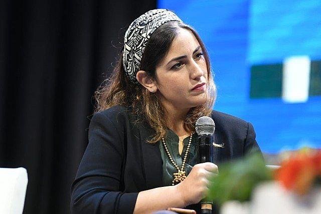 A woman wearing a black-and-white patterned headband and a dark blazer holds a microphone while seated at what appears to be a panel or conference.