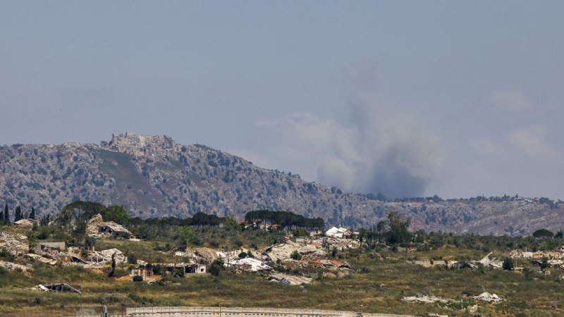 Smoke rises from a forested hillside behind a rural settlement with scattered houses and trees under a clear sky.