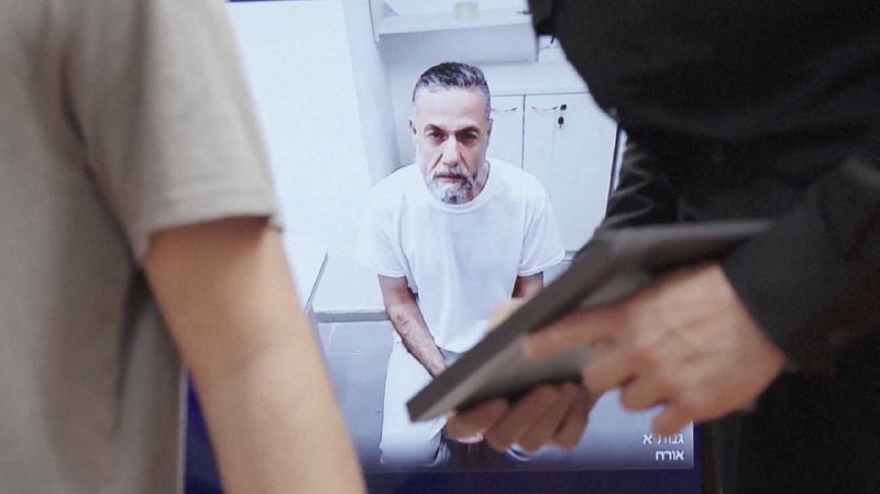 A gray-haired, bearded man in a white shirt sits in a white room, appearing on a screen held by a person in the foreground.