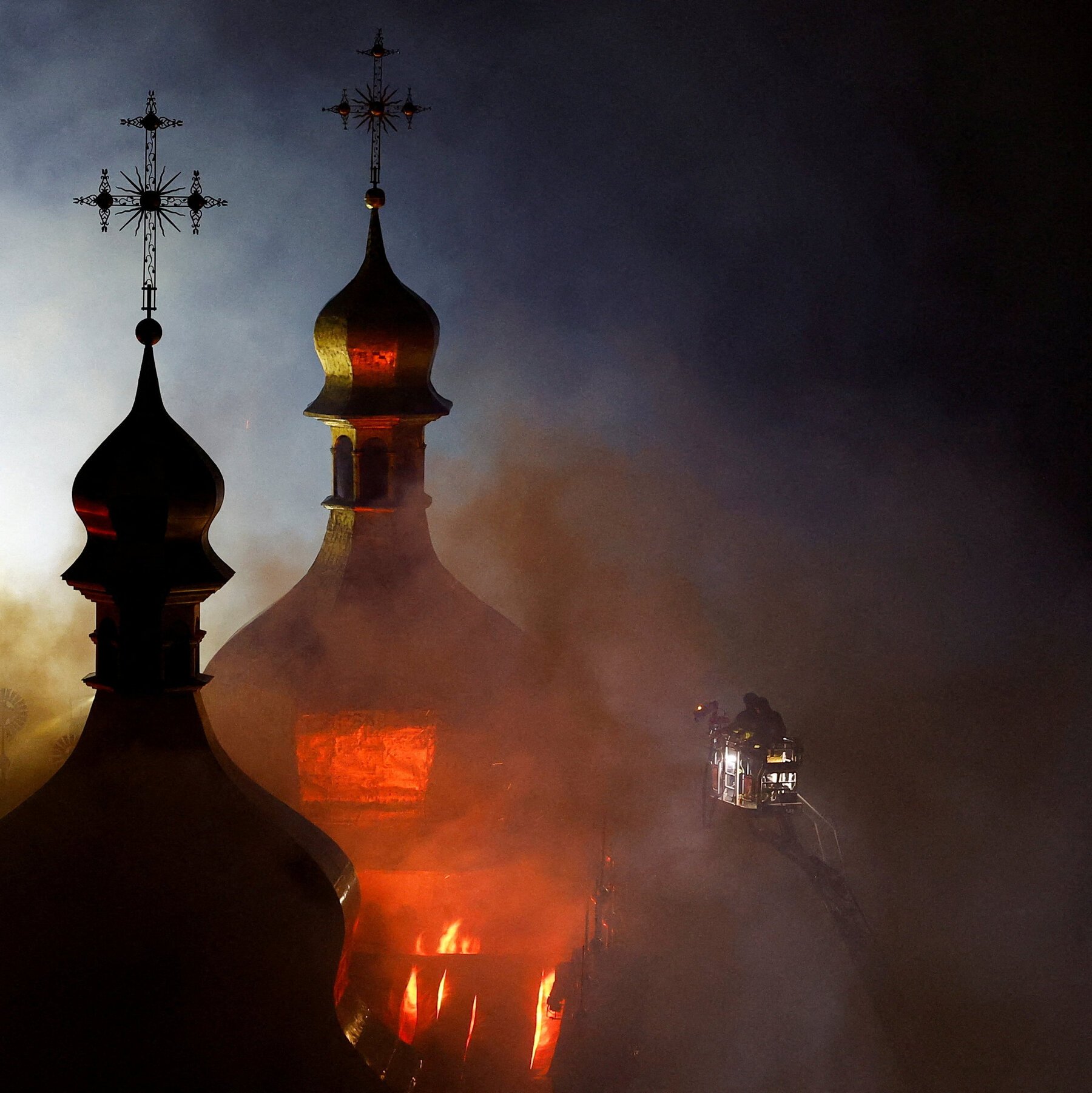 Firefighters in a ladder truck battle a blaze engulfing a church with two onion domes topped by crosses at night.