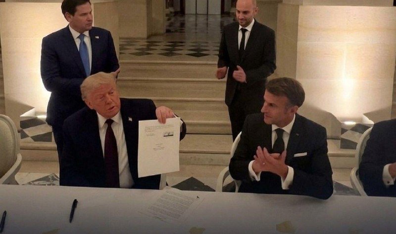 Four men in suits sit and stand around a table in a formal room, with one seated man holding up a document displaying an image of a ship.