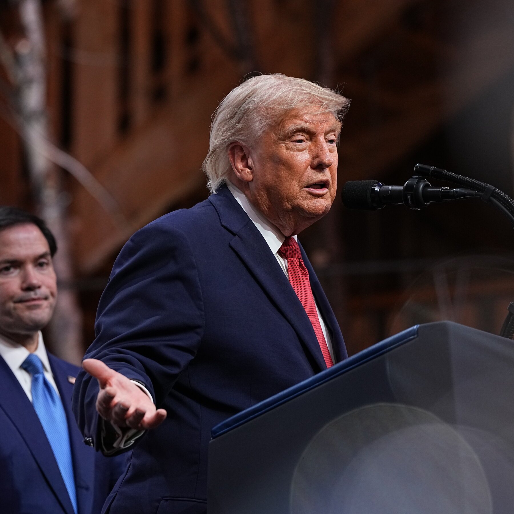 A man with light blonde hair in a navy suit and red tie speaks at a podium with microphones, gesturing with his hand, while another suited man stands behind him.