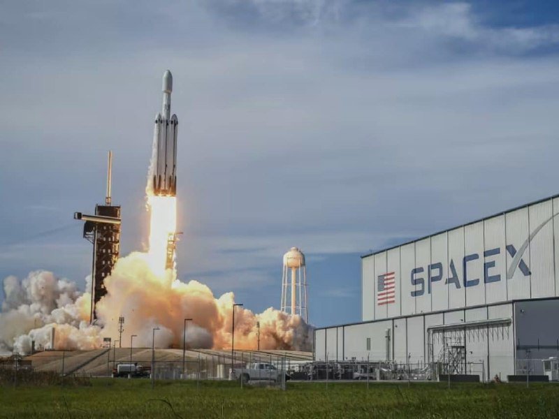 A rocket lifts off from a launch pad, emitting flames and large plumes of smoke, with a SpaceX building and American flag visible in the background.