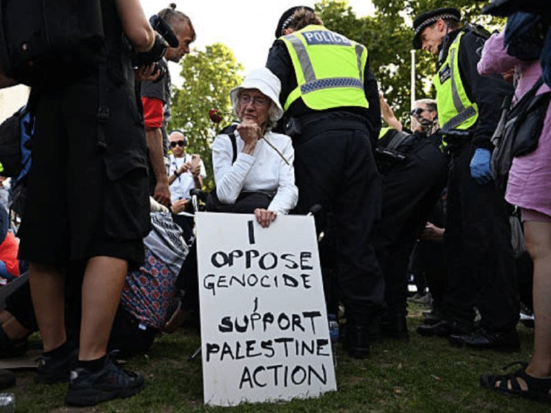 An older seated protester holds a handwritten sign reading "I OPPOSE GENOCIDE, SUPPORT PALESTINE ACTION" while flanked by police officers in high-visibility vests and a crowd.