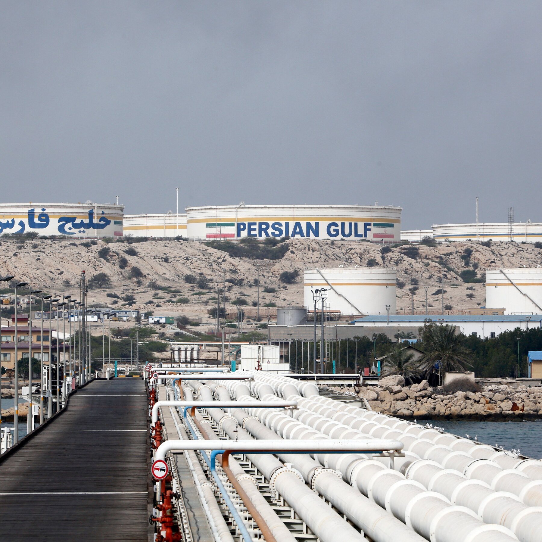White pipelines stretch across a pier toward large storage tanks labeled "PERSIAN GULF" alongside Arabic script and an Iranian flag, set against a rocky shoreline.