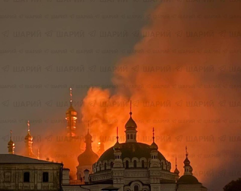 A large fire with bright orange flames and thick smoke rises behind the silhouettes of church domes and spires at night, watermarked with repeating "ШАРІЙ" text.