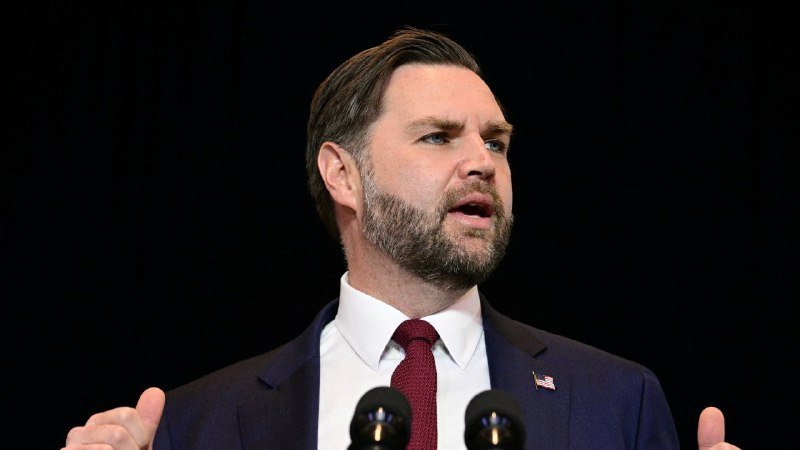 A bearded man in a navy suit, white shirt, red tie, and American flag lapel pin speaks at a podium with microphones against a dark background.