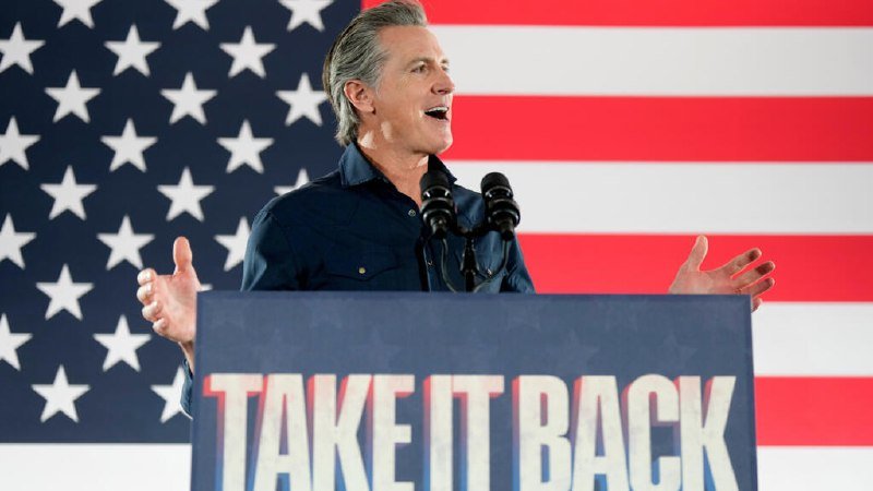 A gray-haired man in a dark shirt speaks at a podium reading "TAKE IT BACK," gesturing with open hands before a large American flag backdrop.