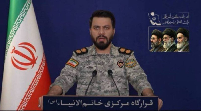 A bearded man in a camouflage military uniform stands at a podium with microphones, with the Iranian flag to his left and Persian text/insignia displayed on the blue backdrop.