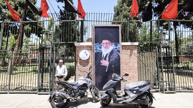 A man stands near two parked motorcycles in front of a brick gate displaying a large framed portrait of a bearded cleric in black robes, with red and black flags hanging above.