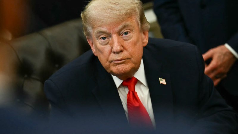 A man with blonde hair wearing a dark suit, white shirt, red tie, and an American flag lapel pin sits with a serious expression in an indoor setting.