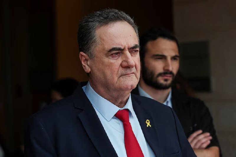 An older man in a dark suit and red tie with a yellow ribbon pin stands with a serious expression, while a younger bearded man stands behind him with crossed arms indoors.