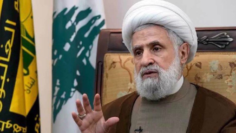 Hezbollah Secretary General Naim Qassem, wearing a white turban and brown robes, gestures with his hand while seated in front of the Lebanese flag and Hezbollah emblem.