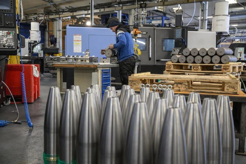 A worker in a beanie operates machinery in an industrial facility, with rows of metallic cone-shaped objects arranged in the foreground near wooden pallets.