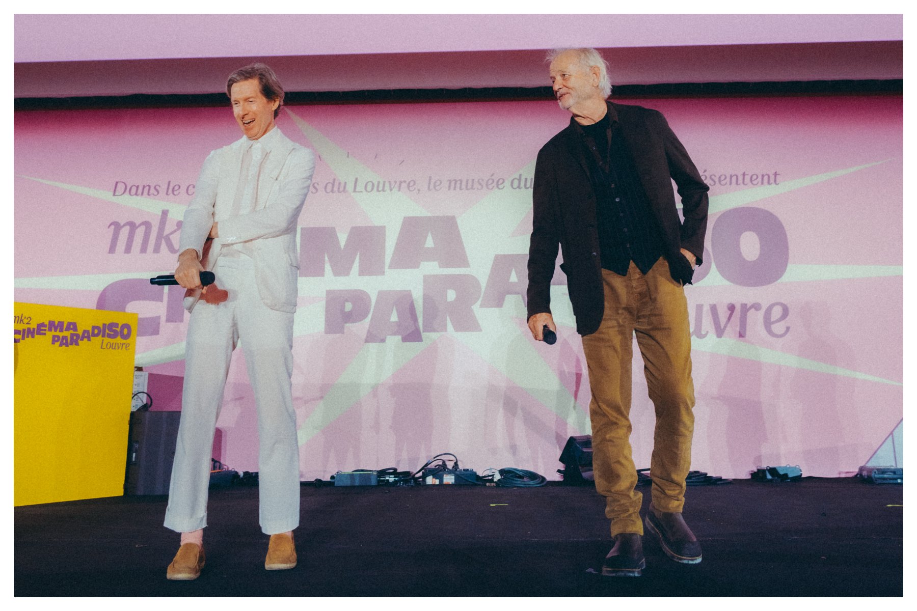 Two men, one in a white suit holding a microphone and another in a dark blazer with tan pants, stand smiling on a stage in front of a "MK2 Cinéma Paradiso Louvre" backdrop.