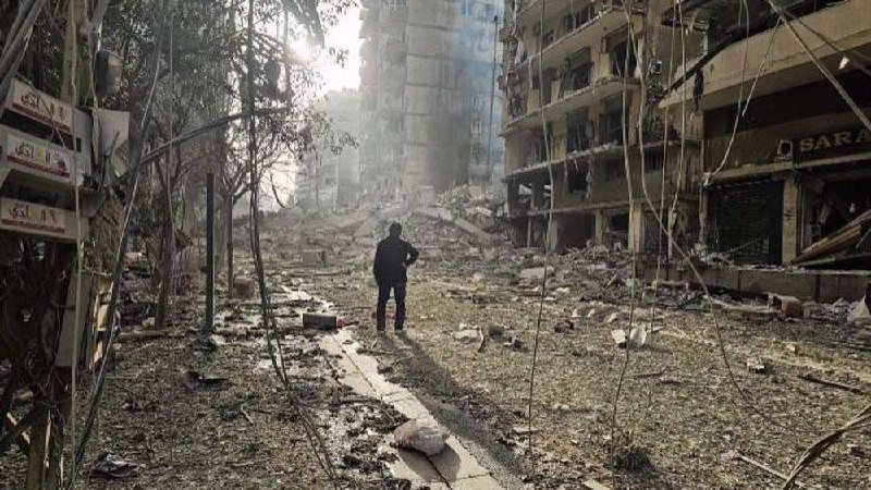 A person stands amid rubble-strewn streets between heavily damaged multi-story buildings, surveying the destruction under hazy, smoke-filled skies.