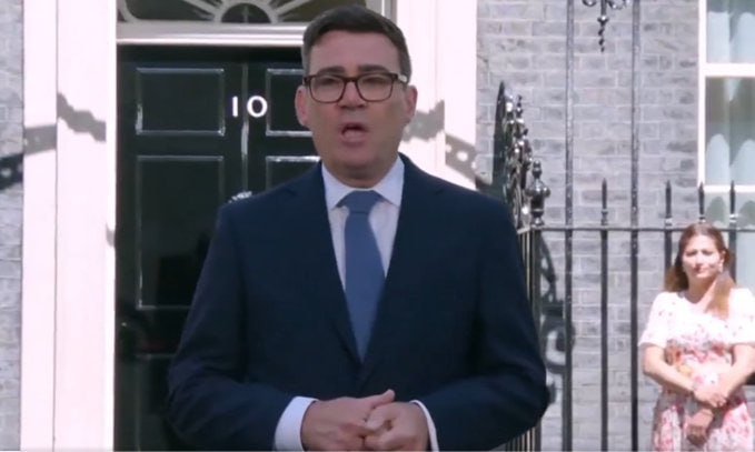 A bespectacled man in a dark suit and blue tie stands speaking in front of a black door marked "10," with a woman in a floral dress standing behind a wrought-iron gate.