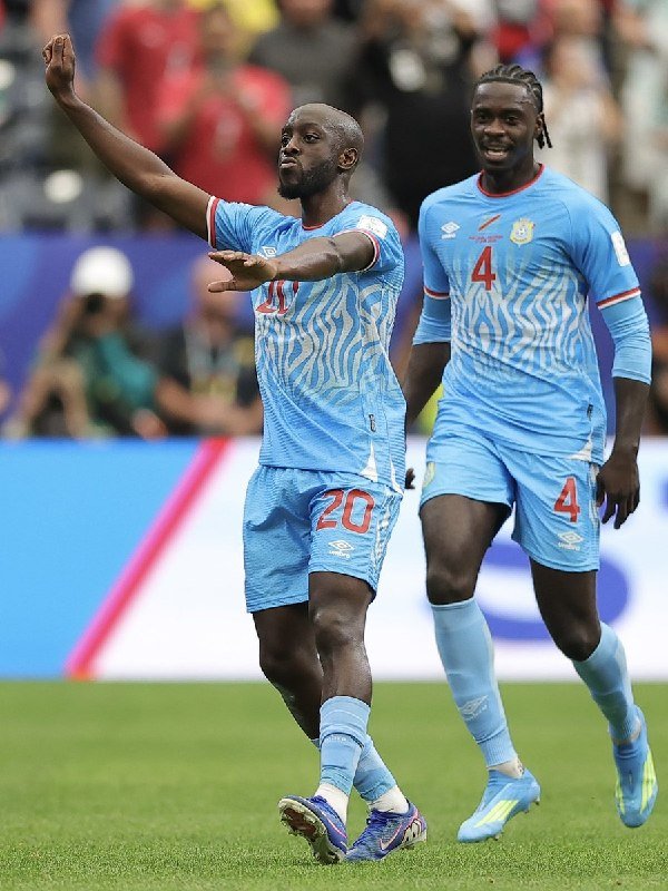 Two soccer players in light blue jerseys with red numbers 20 and 4 celebrate on a grass field, with one raising his arm toward a blurred crowd.