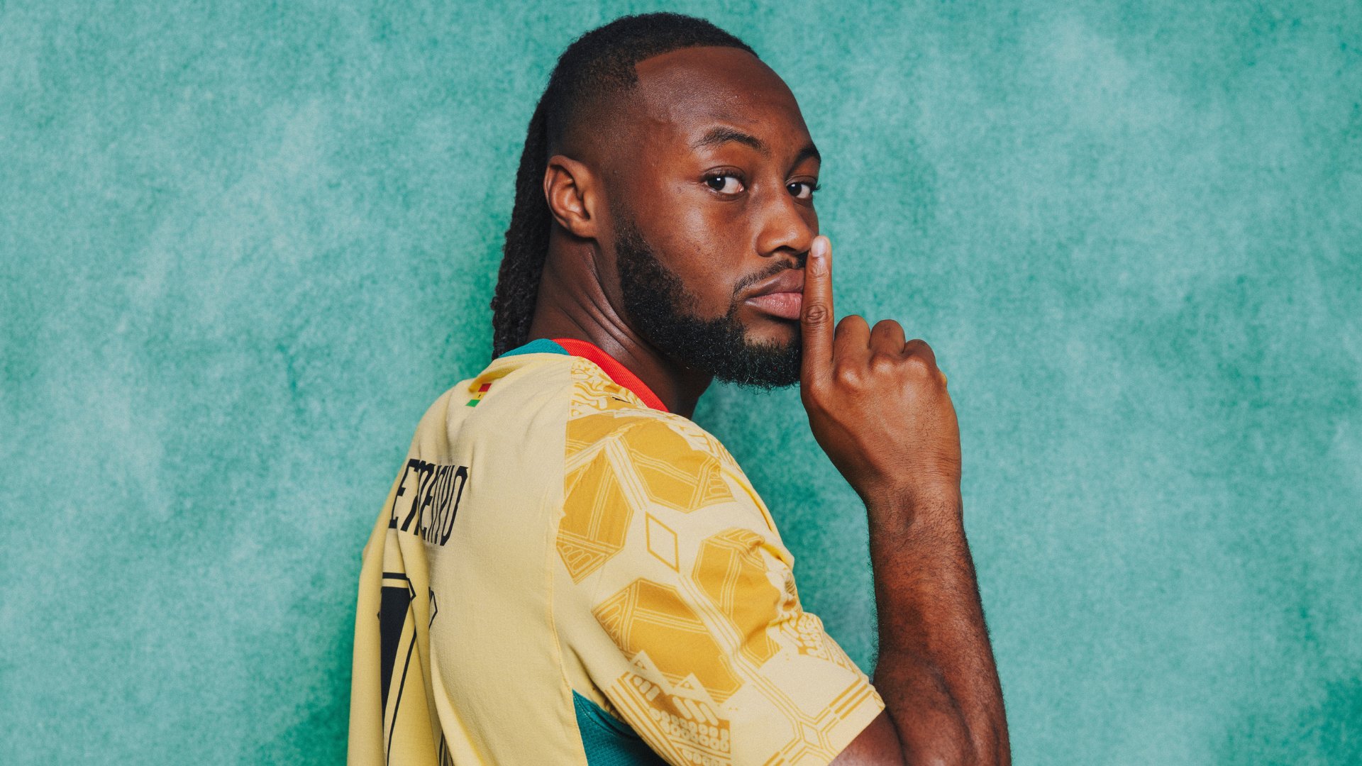 A man with dreadlocks wearing a yellow jersey with the name "AYEW" visible stands against a teal background, holding his index finger to his lips.