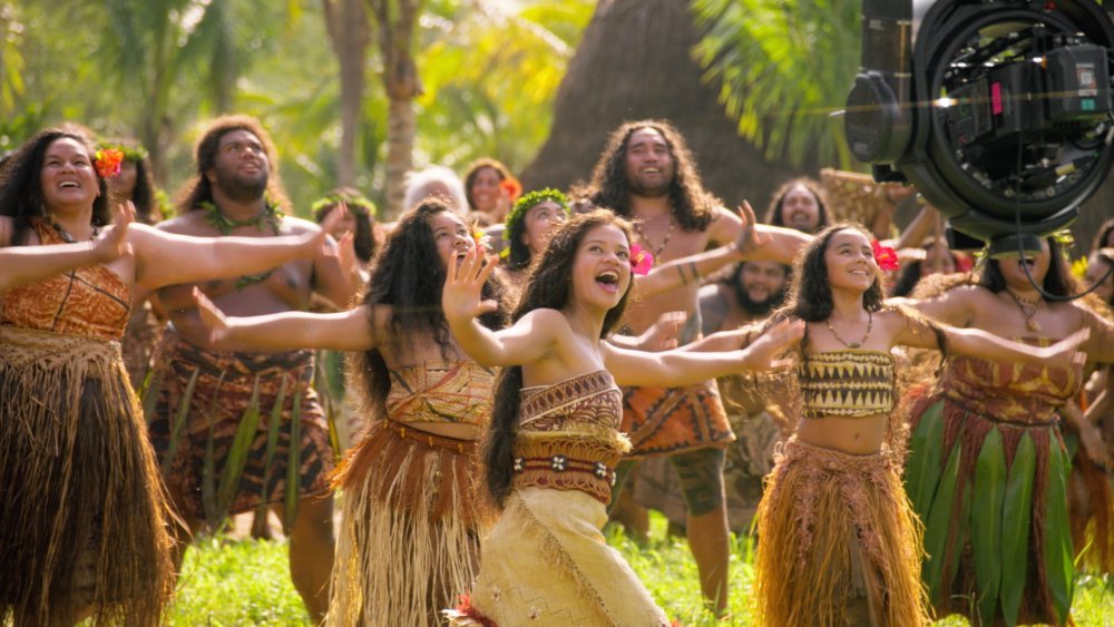 A film camera captures a group of people in traditional tropical attire performing a hula dance outdoors among palm trees.