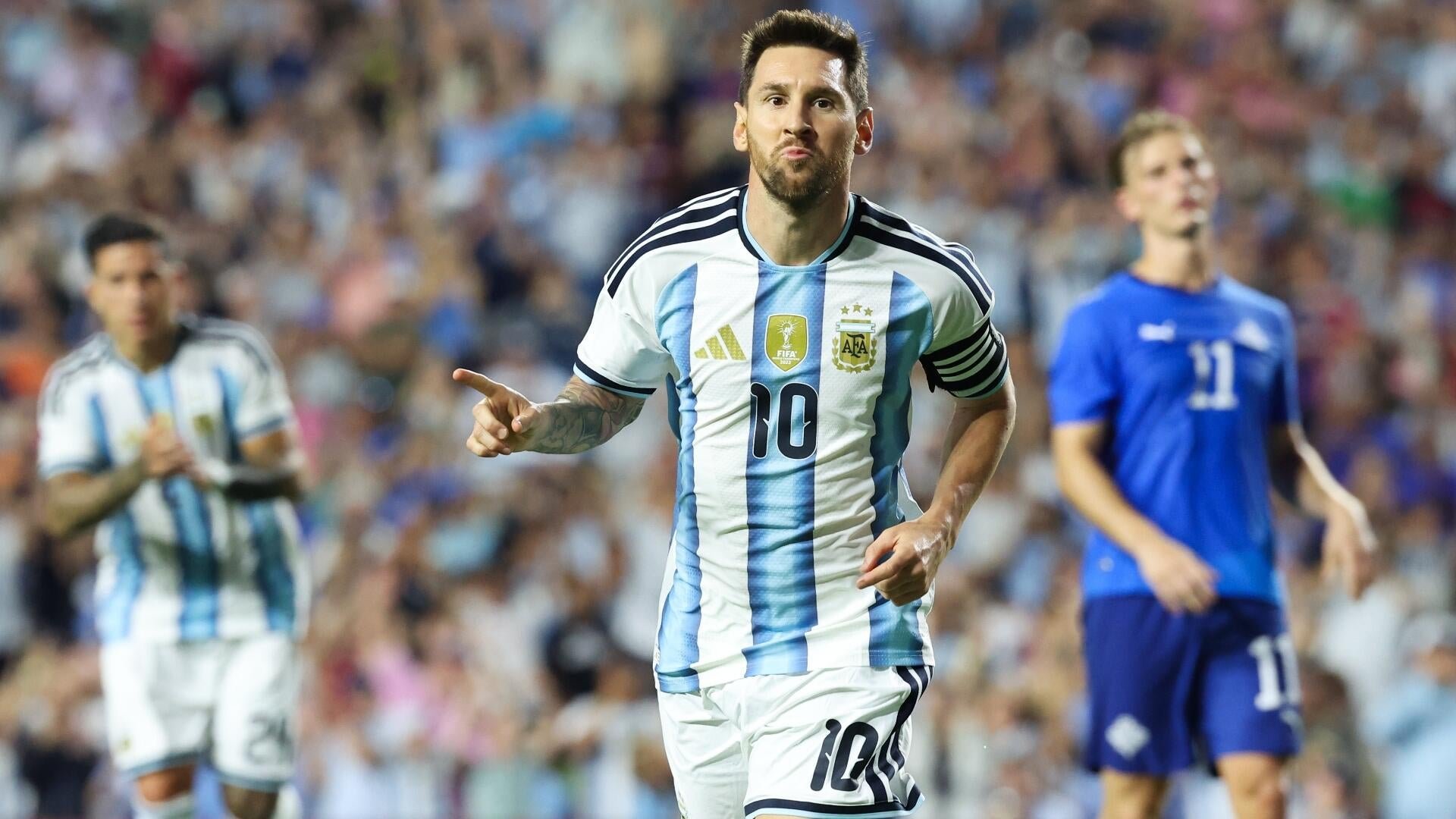 A soccer player wearing a light blue and white striped jersey with the number 10 points forward while running on a field, with a crowded stadium in the background.