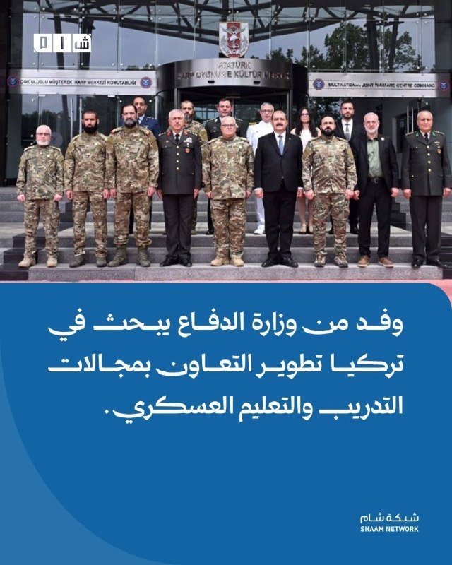 A group of military personnel in uniform pose for a photo on the steps of a building labeled "Multinational Joint Warfare Centre Command," accompanied by Arabic text below.