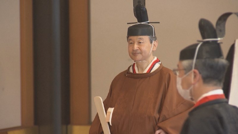 A man in traditional Japanese court attire and a tall black eboshi hat stands holding a wooden tablet, with another masked official visible nearby.