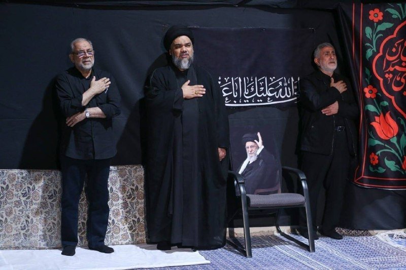 Three men stand with hands over their hearts on a stage with black banners featuring Arabic calligraphy and a portrait of a seated cleric in a turban.