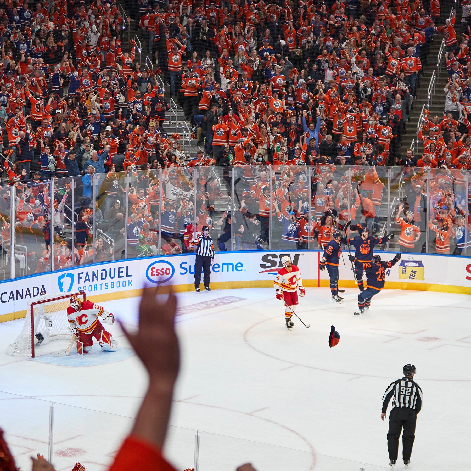 Fans in orange jerseys cheer with raised pom-poms as dark-jerseyed hockey players raise their sticks in celebration on the ice.