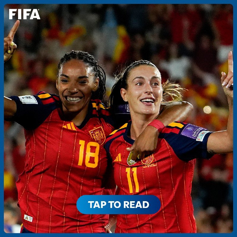 Two smiling female soccer players in red Spain jerseys with numbers 18 and 11 celebrate together with raised arms in a crowded stadium, with a "TAP TO READ" button overlaid.