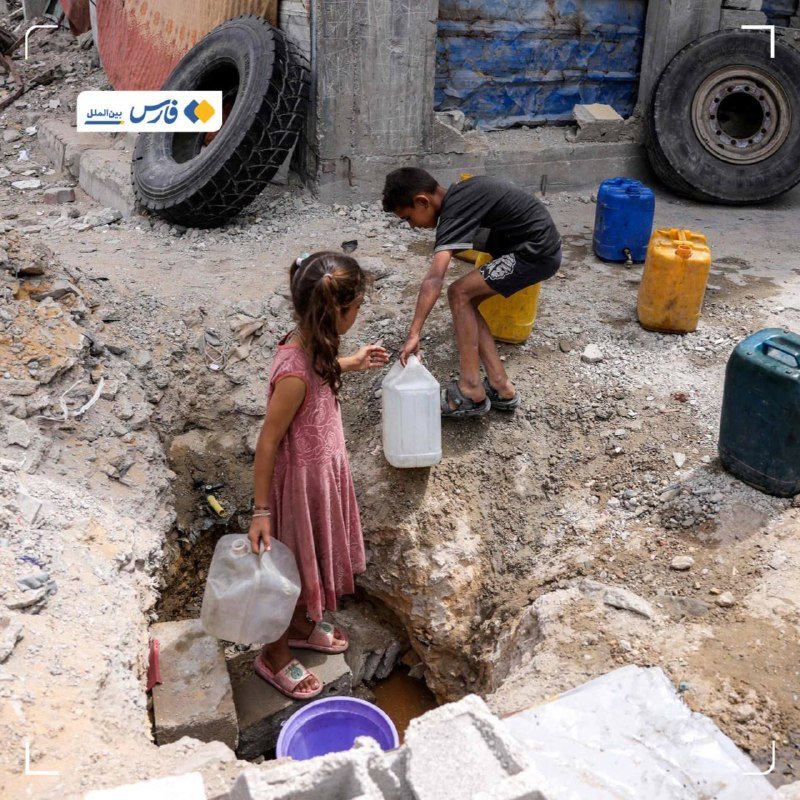 Two children collect water from a muddy, rubble-filled street using plastic jugs and containers beside large tires and a weathered building.