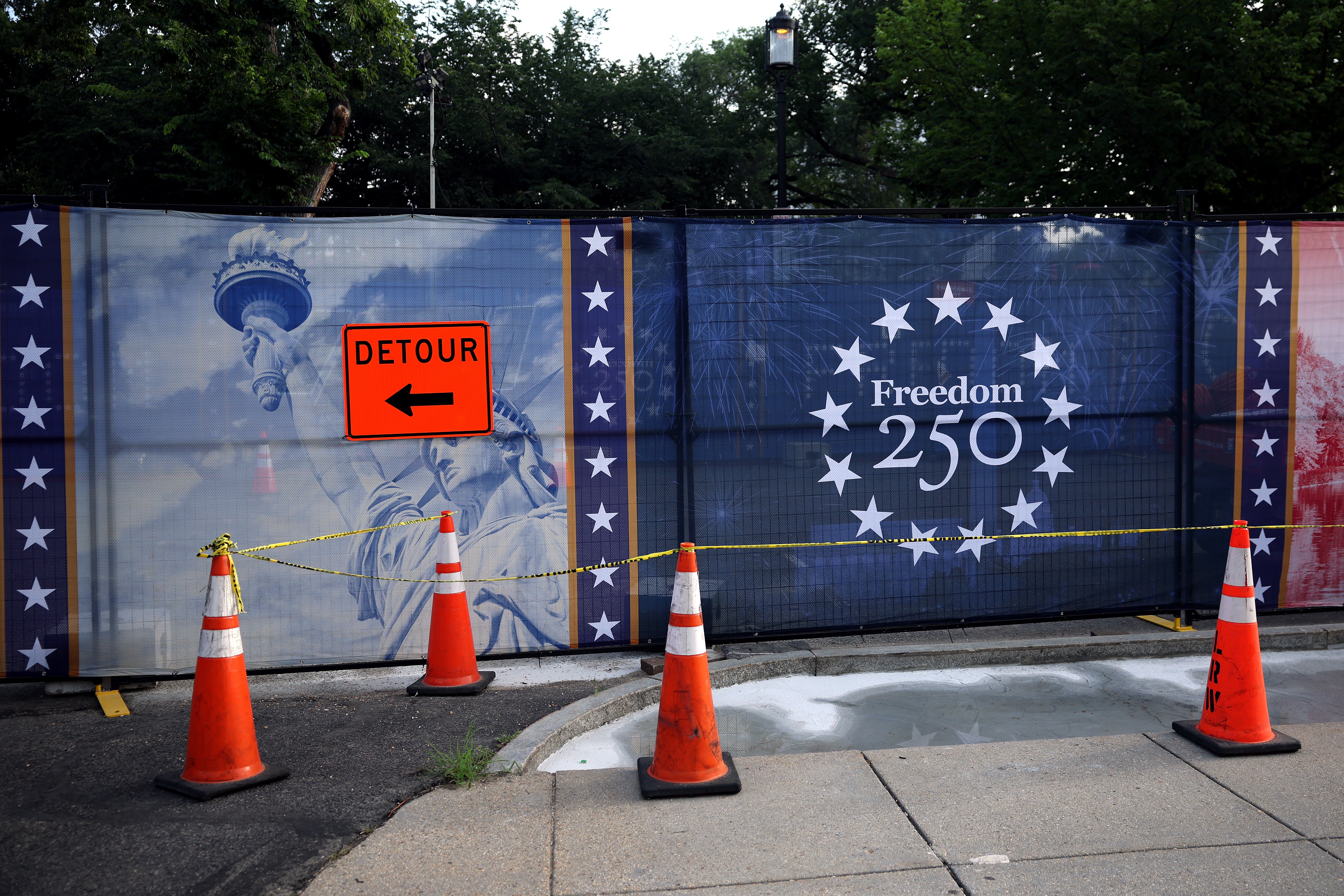 Orange traffic cones, caution tape, and a "Detour" sign block a sidewalk in front of a fence displaying patriotic "Freedom 250" banners.