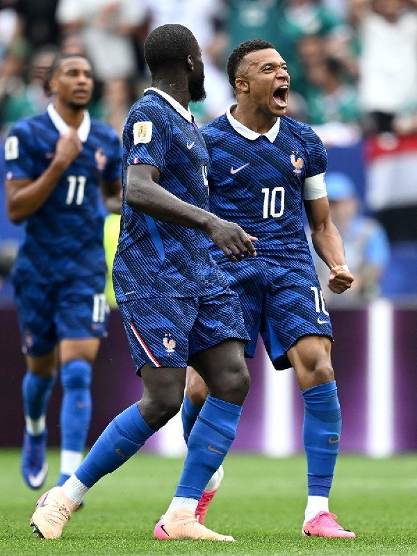 Soccer players in blue France jerseys, including numbers 10 and 11, celebrate on a grass pitch with a roaring shout and clenched fist during a match.
