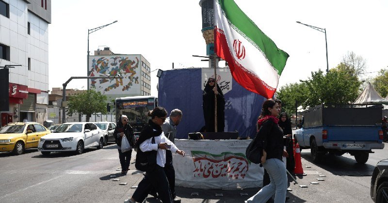 A woman wearing a chador stands on a platform waving a large Iranian flag on a city street, while pedestrians and vehicles pass by.
