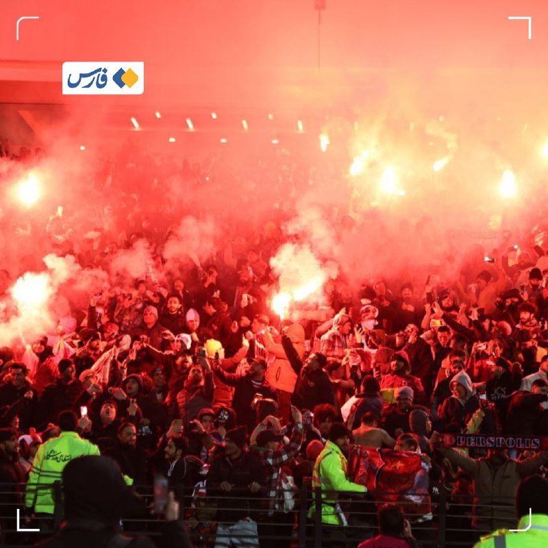 A large crowd in a stadium sets off red flares, filling the air with thick red smoke while several people hold up smartphones to record the scene.