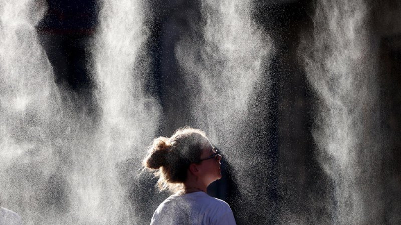 A woman with her hair in a bun stands amid a large spray of illuminated water mist.