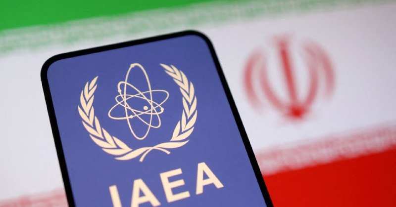 A smartphone displays the IAEA logo and acronym in front of the blurred Iranian flag.
