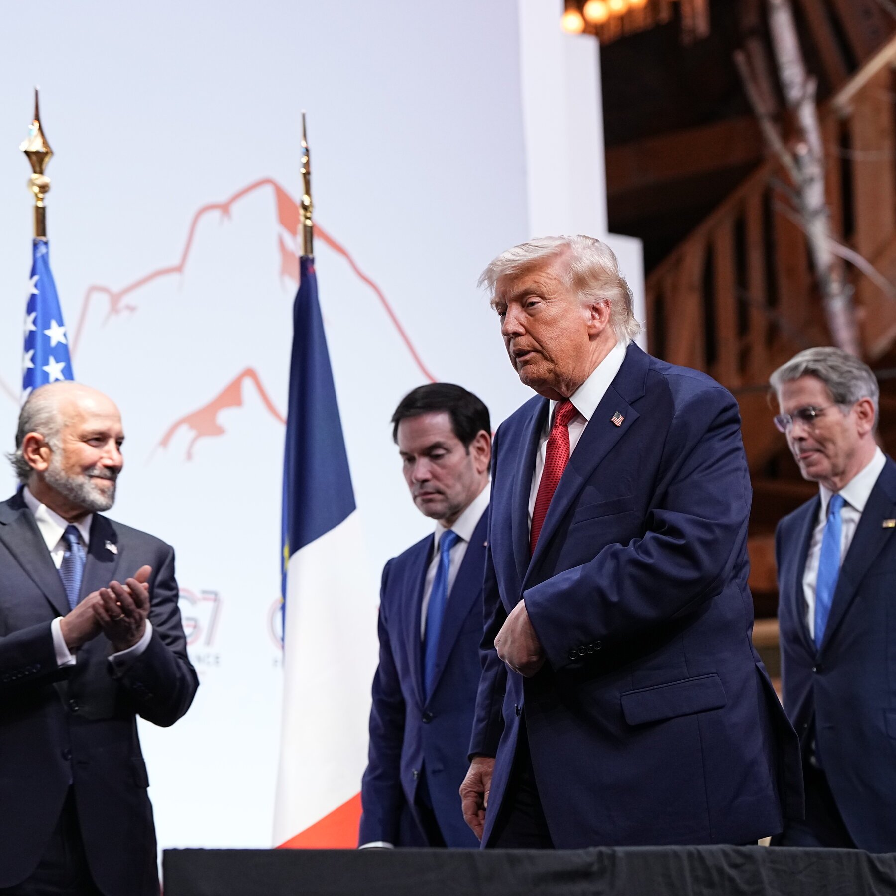A white-haired man in a navy suit with a red tie walks past three other suited men, with U.S. and other flags visible in the background.