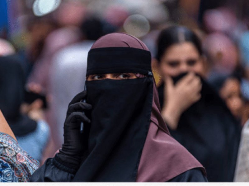 A woman wearing a black niqab and gloves holds a phone to her ear in a crowded setting, with other people visible behind her.