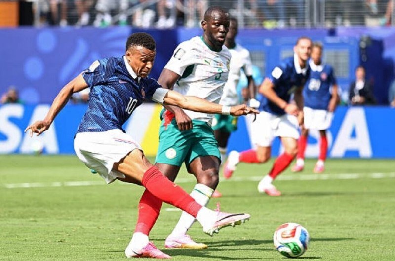 A soccer player in a blue jersey kicks the ball while being challenged by an opponent in a white and green uniform on a stadium field.