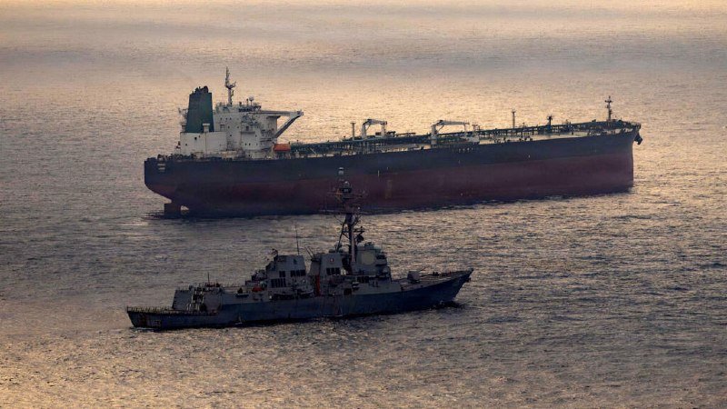 A small naval warship sails past a large tanker ship on calm open water at dusk.