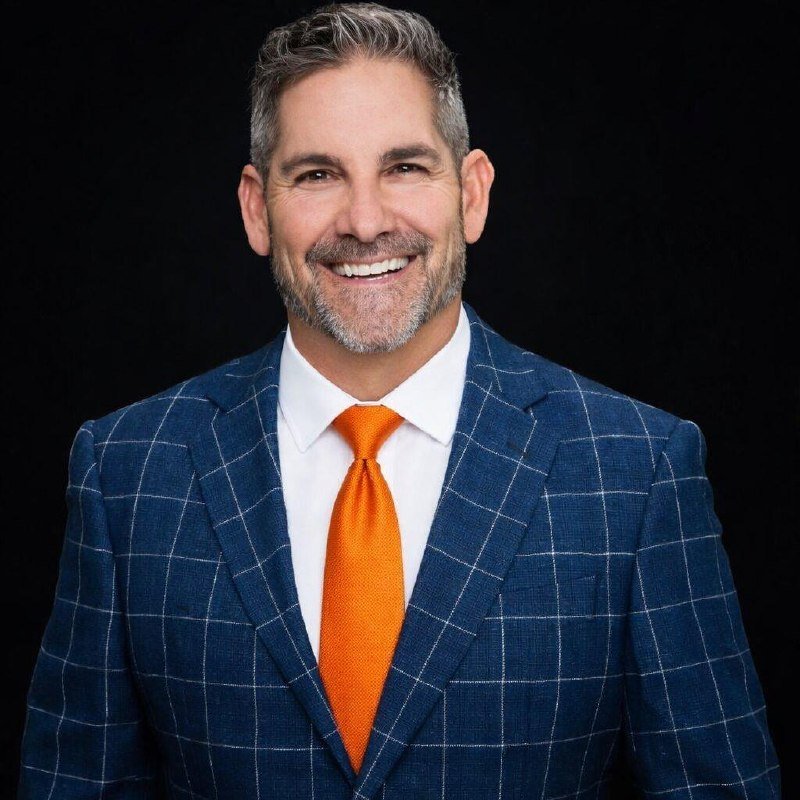 A smiling man with graying hair and beard wears a blue windowpane-check suit, white shirt, and bright orange tie against a black background.