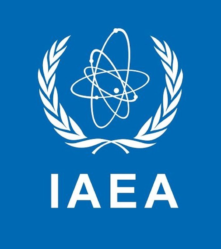 The IAEA logo, featuring a white atom symbol encircled by laurel branches above the acronym "IAEA," appears on a blue background.
