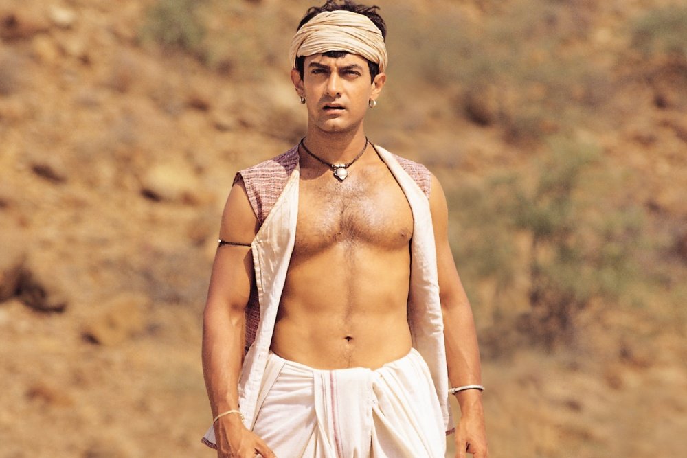 A shirtless man wearing a head wrap, open vest, white dhoti, and pendant necklace stands against a rocky, arid hillside.