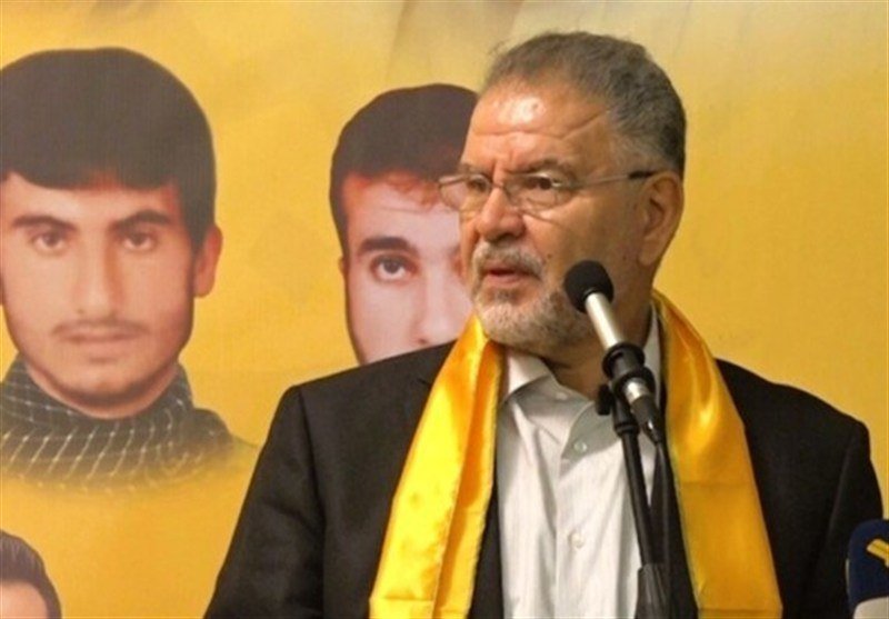 An older bearded man wearing glasses and a yellow sash speaks into a microphone, with large portraits of two younger men displayed on a yellow backdrop behind him.