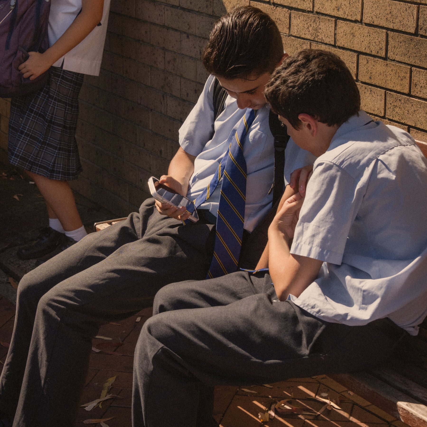 Two boys in school uniforms sit on a bench against a brick wall, one looking at a smartphone while the other leans in to view the screen.