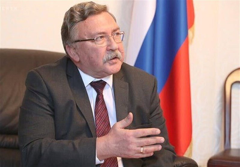 A gray-haired man wearing glasses, a dark suit, and a red striped tie gestures with his hand while seated in front of a white, blue, and red flag.