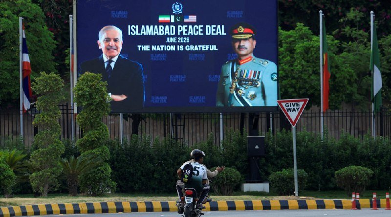 A motorcyclist rides past a large billboard reading "ISLAMABAD PEACE DEAL JUNE 2026" featuring portraits of two officials and flags, with a "GIVE WAY" sign nearby.