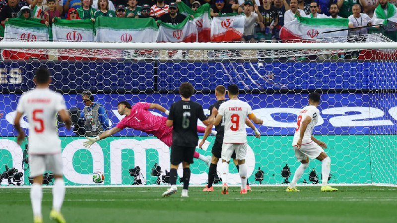A goalkeeper in pink dives toward a ball near the goal line during a soccer match, with players in white and black jerseys watching and fans holding Iranian flags in the stands.